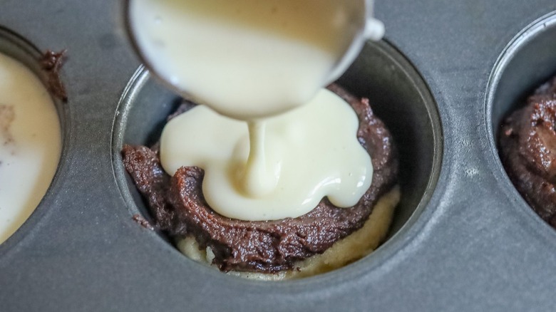 Cheesecake batter pouring into muffin tin well with brookie mixture