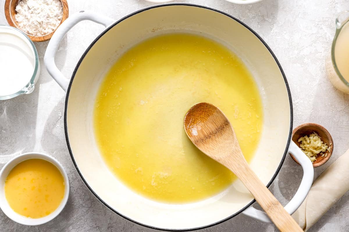 Melted butter and olive oil in a large white skillet with a wooden spoon, with flour, cream, garlic, and egg wash in bowls nearby.
