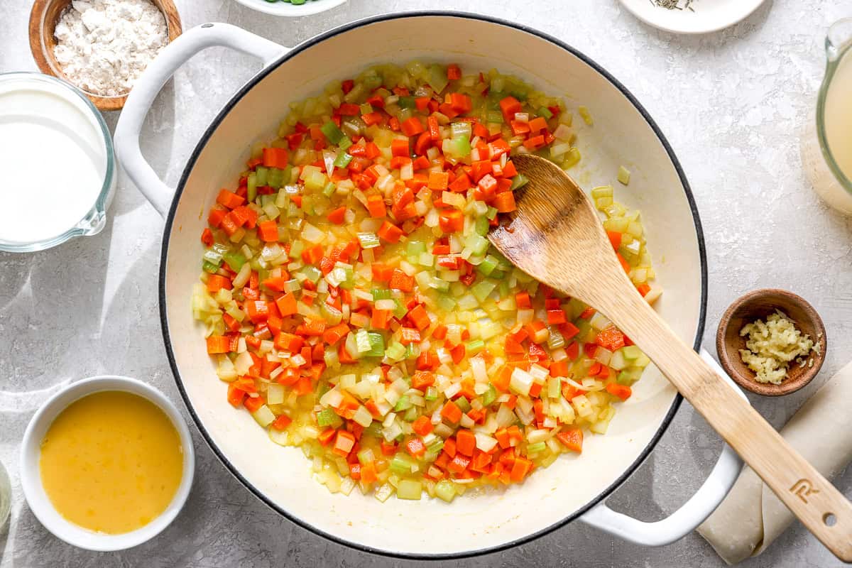 Diced carrots, celery, and onion sautéing in butter in a large white skillet with a wooden spoon.