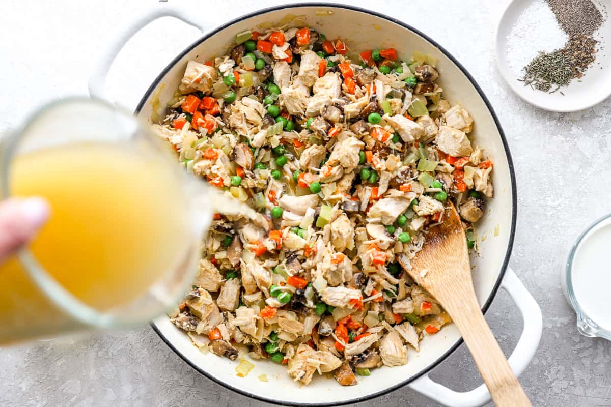Chicken broth being poured into the chicken and vegetable mixture in a large white skillet.