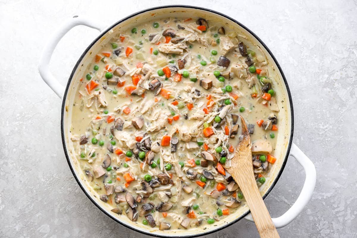 Thickened chicken pot pie filling with shredded chicken, mushrooms, peas, and carrots in a creamy sauce, stirred with a wooden spoon in a white skillet.