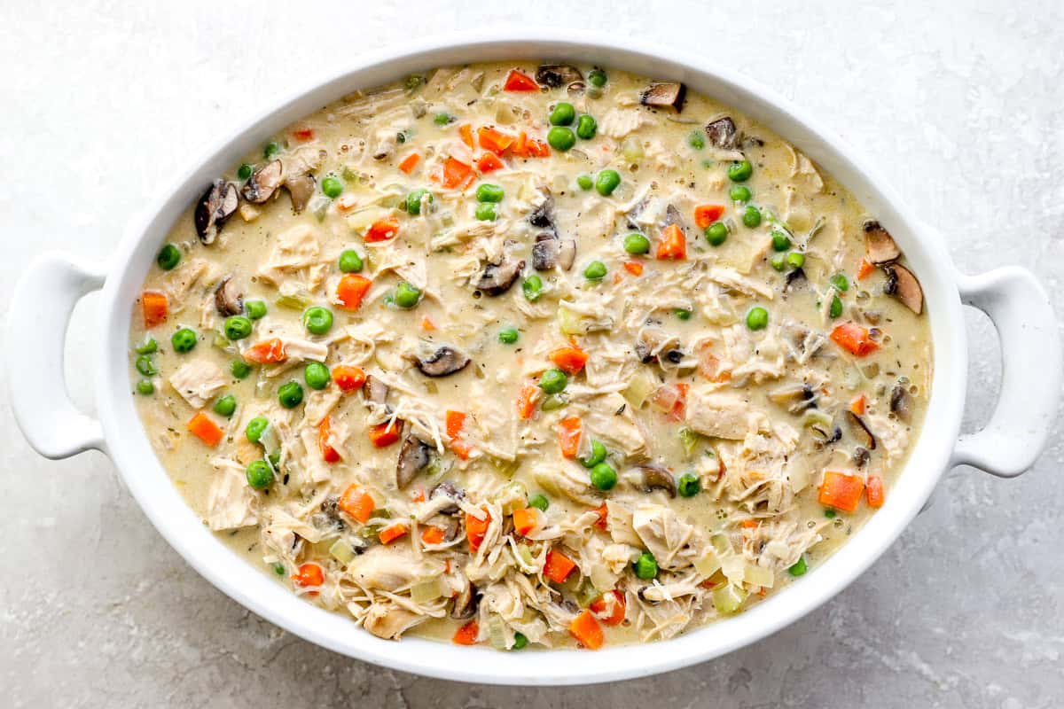 Creamy homemade chicken pot pie filling with chicken, carrots, peas, and mushrooms spread in a white oval baking dish.