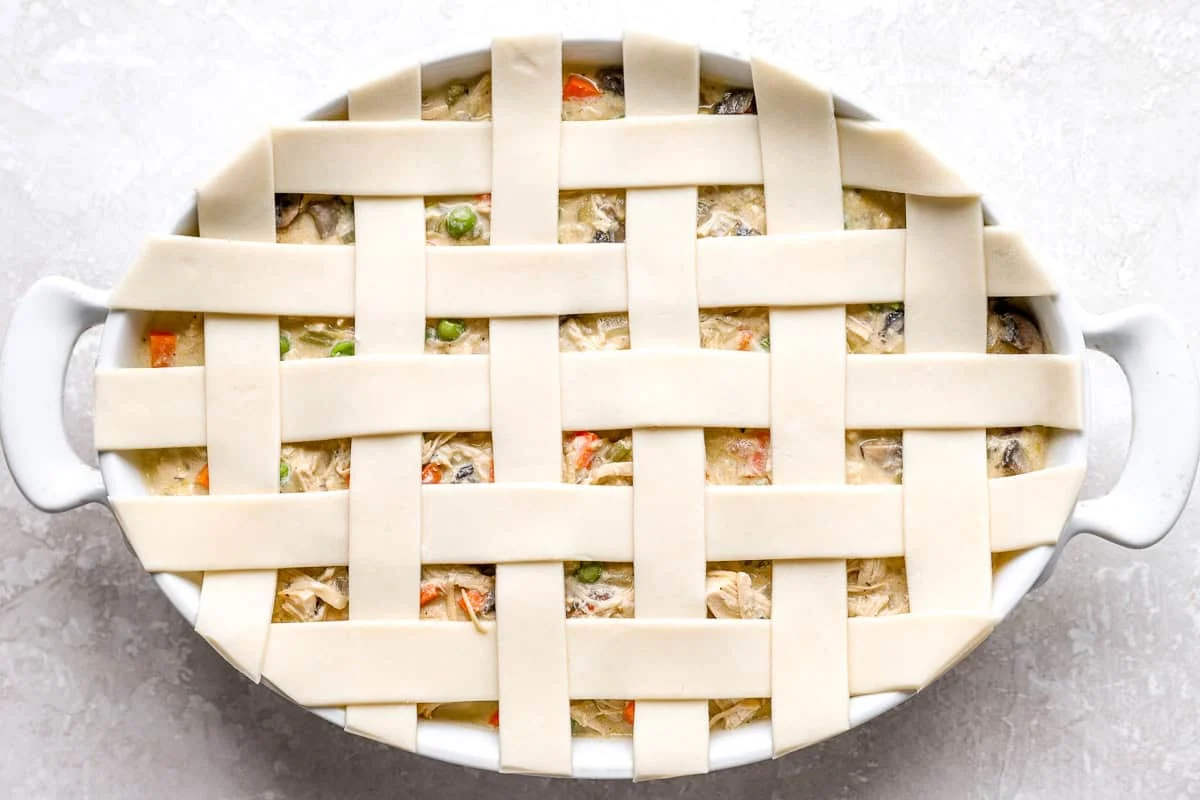Unbaked pie crust strips woven into a lattice over chicken pot pie filling in a white oval baking dish.