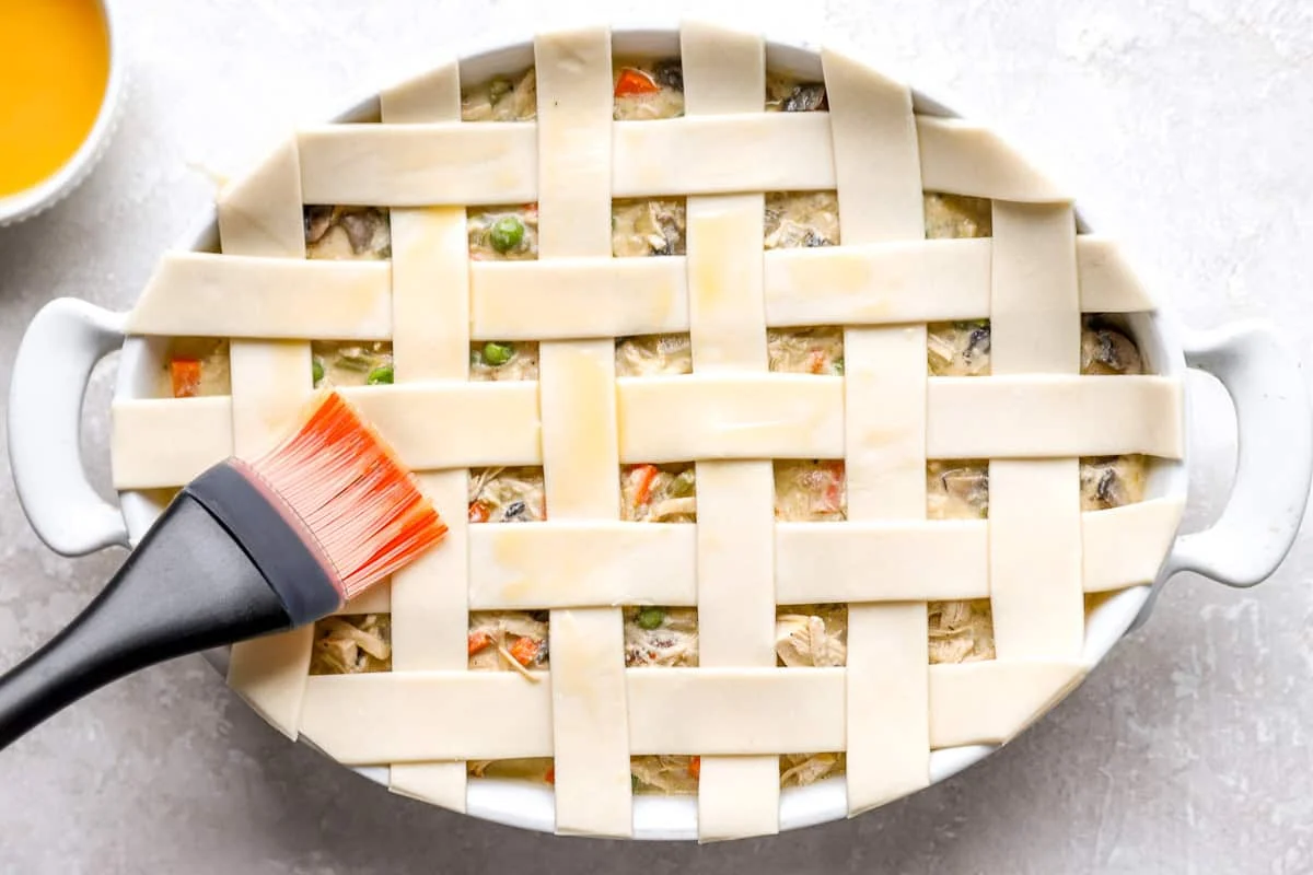 Pastry brush spreading egg wash over the lattice pie crust on an unbaked chicken pot pie casserole.