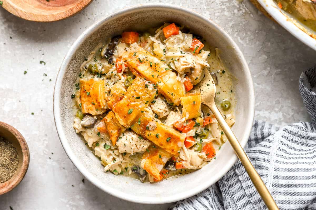 Bowl of creamy chicken pot pie casserole with golden crust pieces, chicken, carrots, and peas, served with a gold fork.
