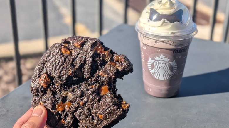 A hand holding a Venti Black Night Cookie with a bite taken out in front of a Black Cat Frappuccino