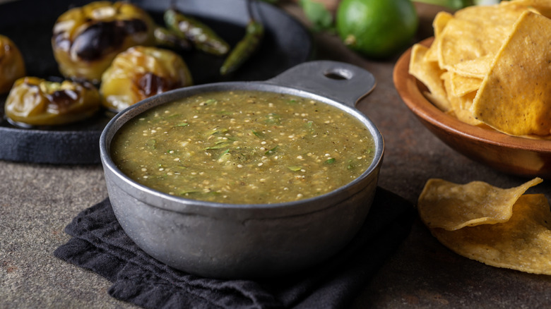 Roasted tomatillo soup beside fresh whole tomatillos and tortilla chips