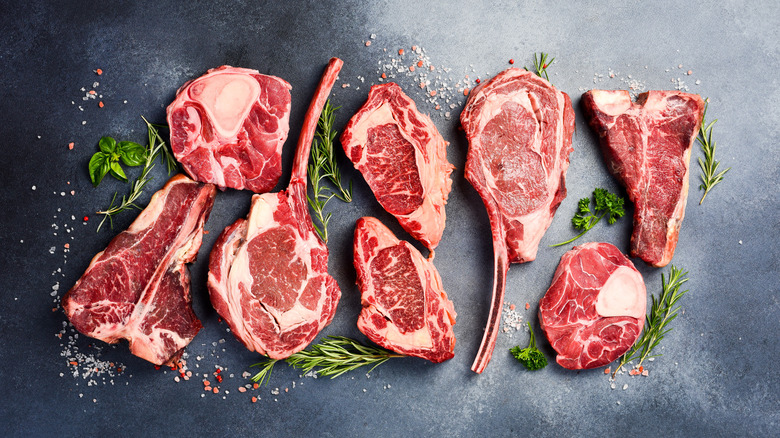 Top-down shot of variety of uncooked steak cuts surrounded by herbs on grey surface