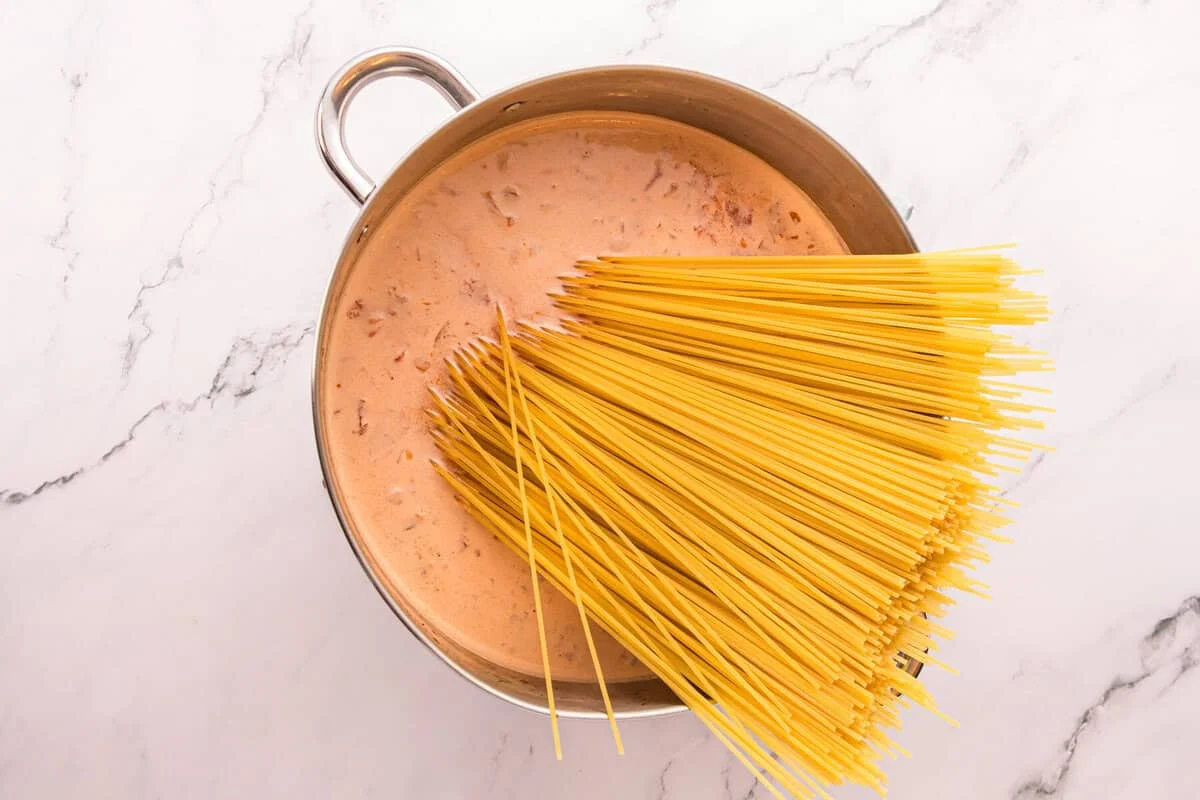 Uncooked spaghetti being added to a creamy tomato broth in a large pot.