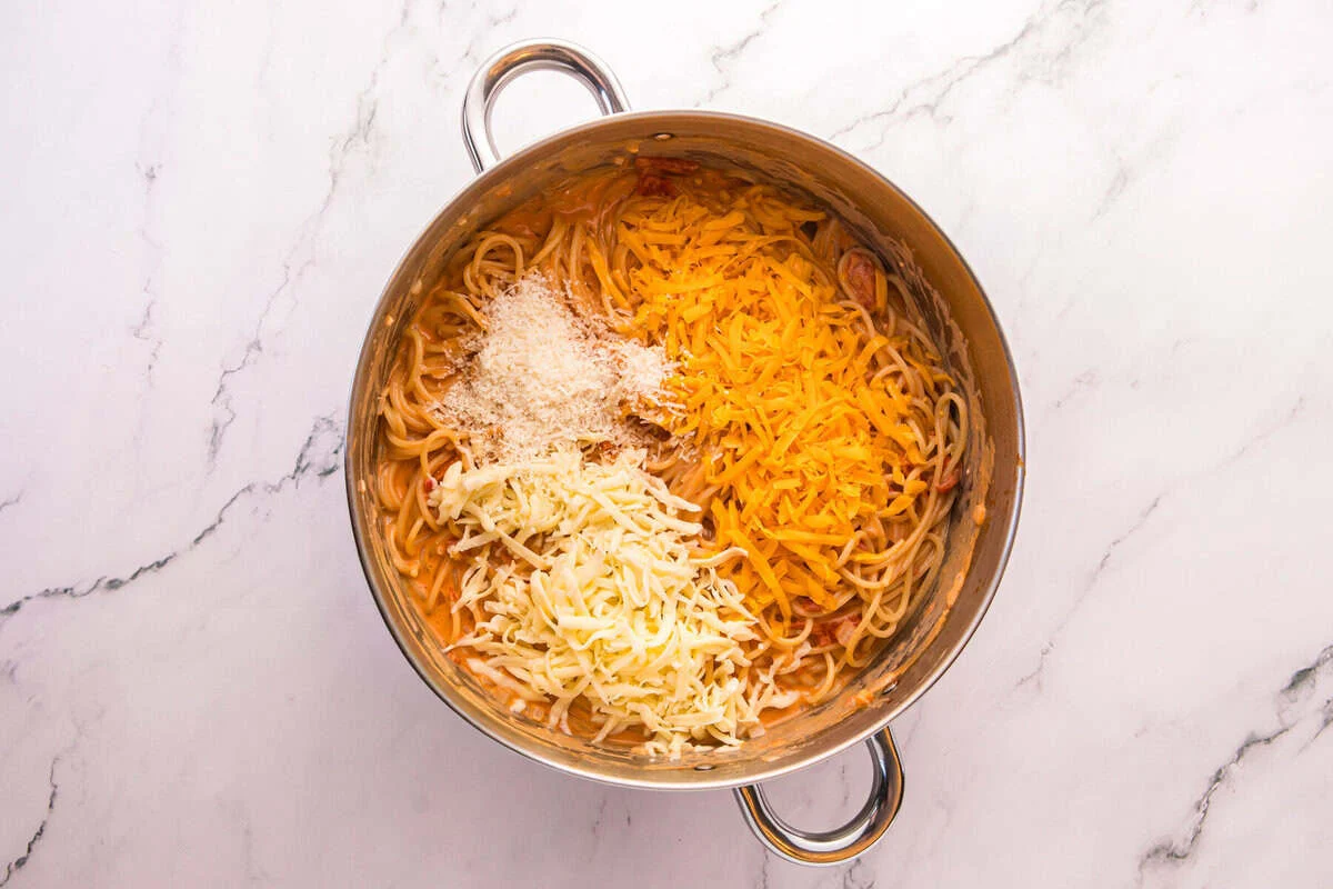 Shredded cheddar, Monterey Jack, and Parmesan cheese added to creamy spaghetti in a pot.