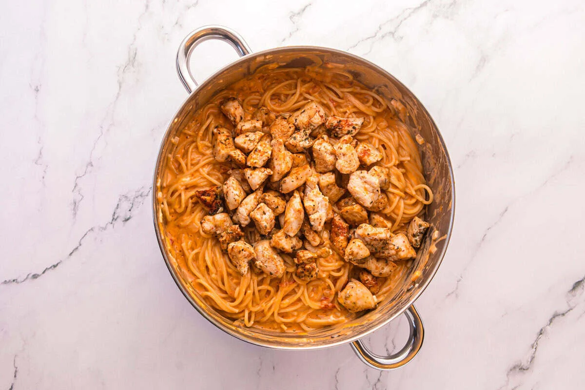 Browned chicken pieces added back on top of cheesy spaghetti in a stainless steel pot.