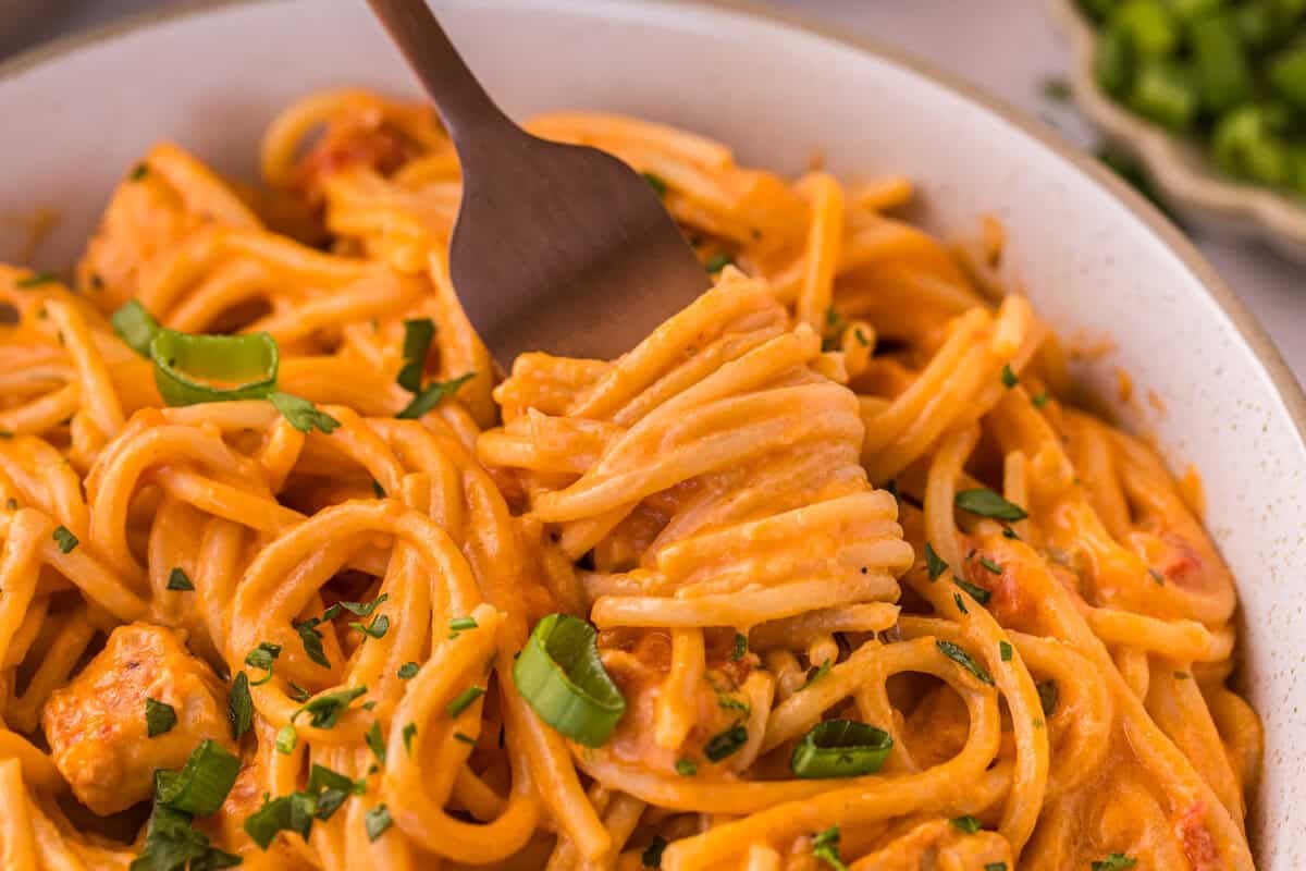 Fork twirling creamy chicken spaghetti in a bowl topped with green onions and parsley.