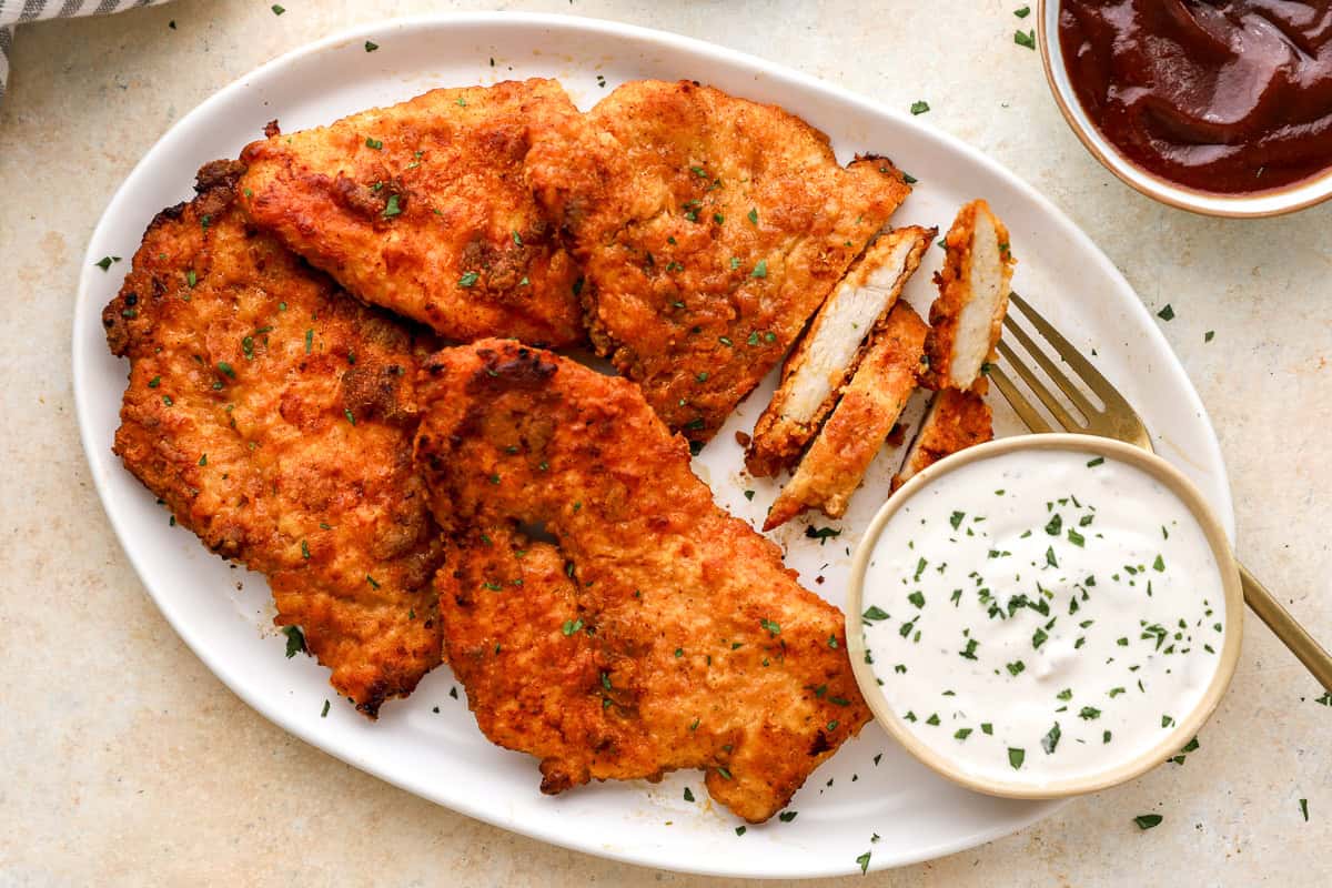 Oven-fried chicken breasts on a platter with one sliced open to show the juicy white meat inside, with ranch and barbecue sauce.