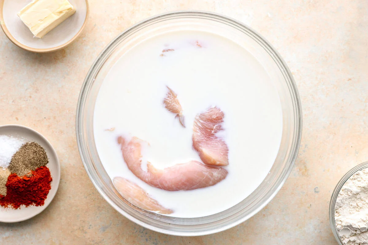 Thin chicken breast cutlets soaking in a glass bowl of milk, with a plate of spices and a bowl of flour nearby.