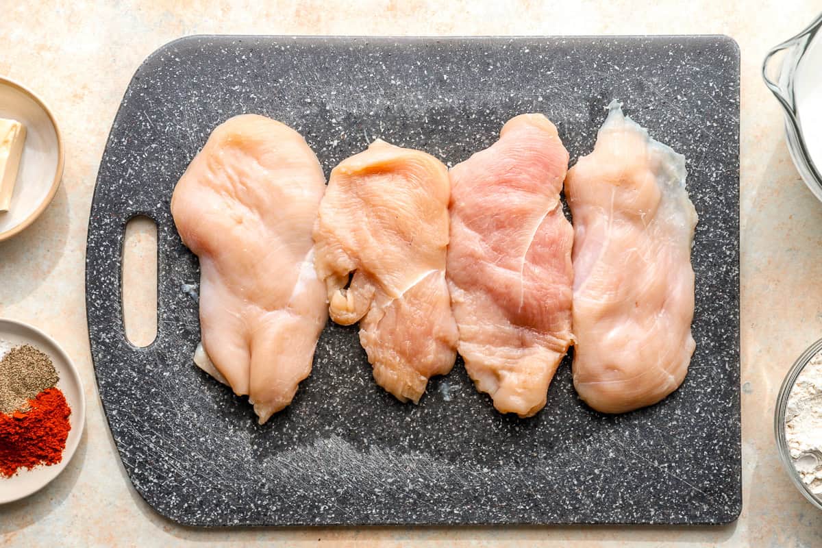 Four thin-sliced raw chicken breasts on a gray cutting board next to small dishes of paprika, black pepper, and butter.