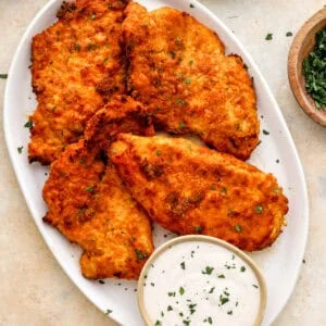 Golden, crispy oven-fried chicken breasts on a white platter with a bowl of ranch dressing and chopped parsley on the side.