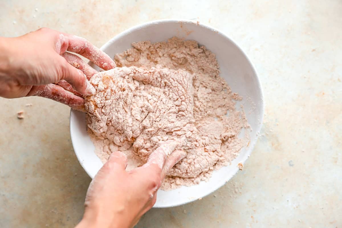 Hands pressing a milk-soaked chicken breast into the seasoned flour to coat it evenly.