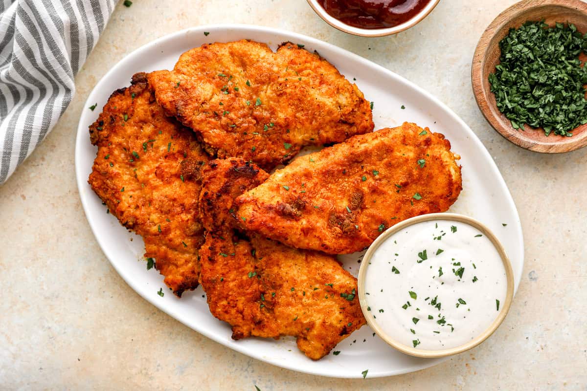 Platter of crispy oven-fried chicken breasts garnished with parsley, served with ranch and barbecue sauce.