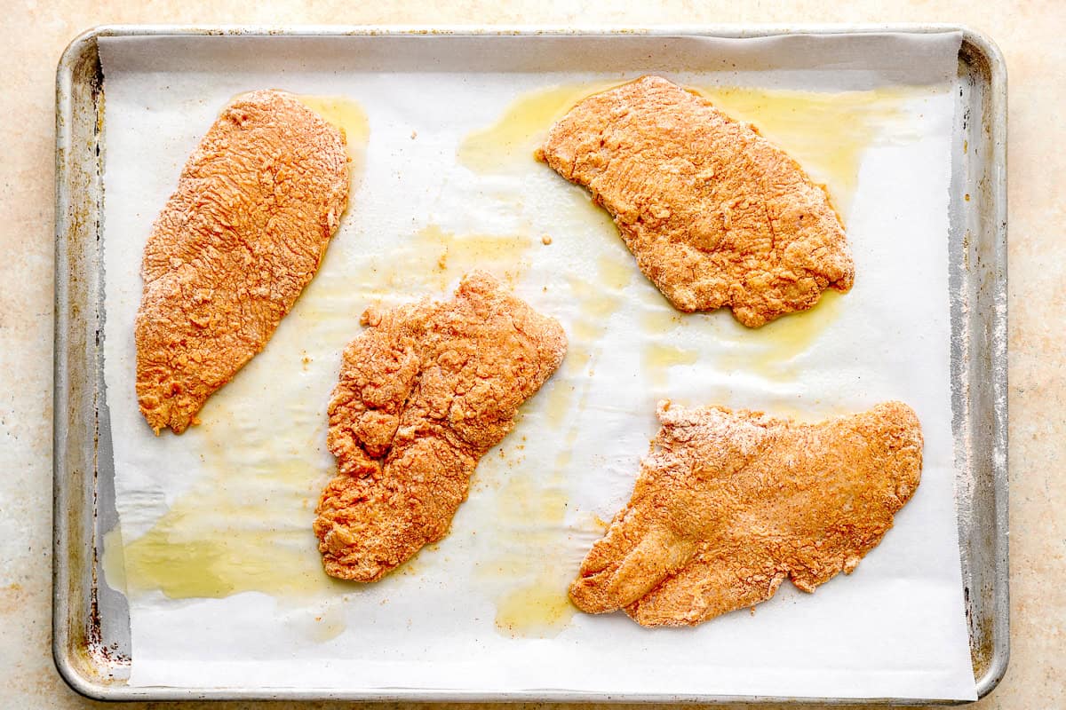 Four flour-coated chicken breasts on a buttered baking sheet, ready to go into the oven.
